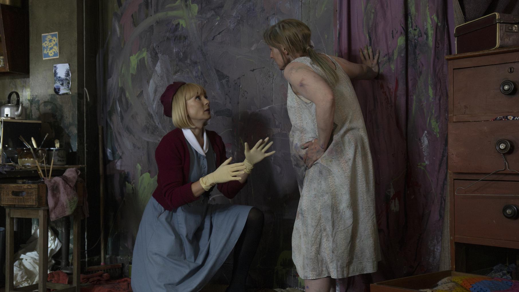 Trixie kneels before a woman in front of a wall daubed with paint